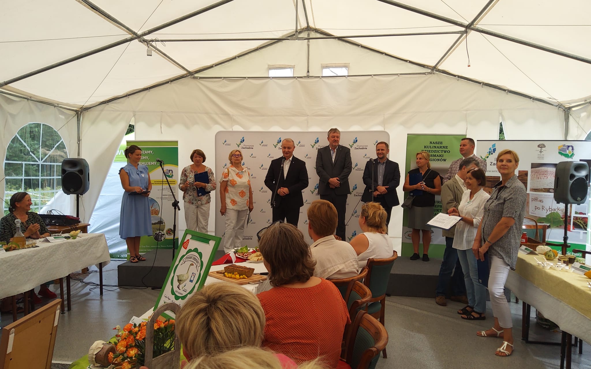 Wnętrze dużego namiotu ogrodowego, w którym znajduje się około 20 osób. Część siedzi przy stołach, osiem osób stoi na podium, trzy - obok. Przy mikrofonach kobieta w niebieskiej sukience i mężczyzna w garniturze. Za nim ścianka z logo Pomorza Zachodniego.