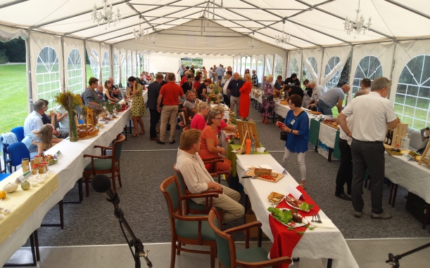 Duży namiot ogrodowy z cześciwowo przezroczystymiu ścianami. W namiocie kilkadziesiąt osób i trzy kilkunastometrowe stoły, na których poukładane są produkty regionalne - słoiki z miodami, wędliny, ciasta, warzywa. 