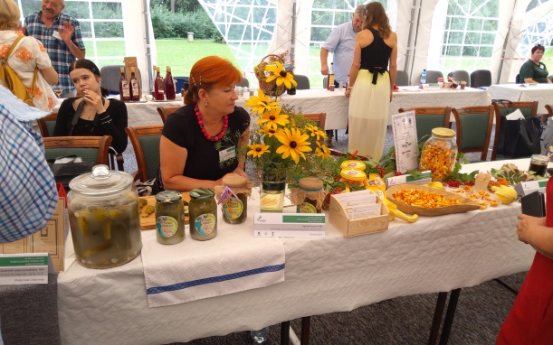 Kobieta w czarnej bluzce i naszyjniku z różowych kul siedzi za stołem, na którym ustawiony jest duży słój z ogórkami kiszonymi, cztery litrowe słoiki i wazon z kwiatami. Obok widać półmisek, słoiki z drobnymi kawałkami dyni, a dalej - dwie stojące osoby.